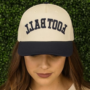May include: A baseball cap with a cream-coloured crown and a navy blue brim. The word "FOOTBALL" is embroidered in navy blue on the front of the cap. The cap is worn by a person.