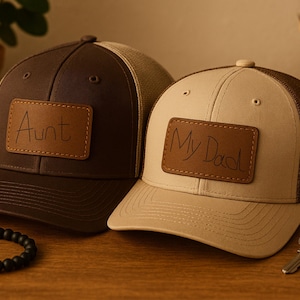 Op de afbeelding: Twee baseballpetten, één bruin en één beige, met mesh-rug. Elke pet heeft een leren patch met de woorden "Aunt" en "My Dad". Een zwarte kralenarmband en sleutels liggen op het houten oppervlak.
