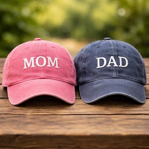 May include: Two baseball caps, one pink and one navy blue, sit side-by-side on a wooden surface. The pink cap has the word "MOM" embroidered in white, while the navy blue cap has "DAD" embroidered in white.