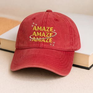 May include: A red baseball cap with a curved brim. The cap features the word "AMAZE" embroidered in yellow, with a stylized design. The hat is made of a soft, textured material and has a classic six-panel design.