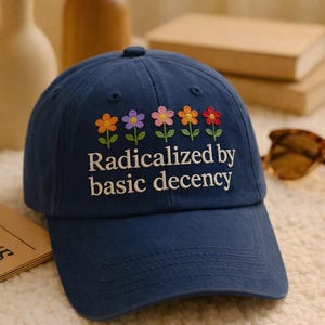 May include: A navy blue baseball cap featuring embroidered flowers in orange, purple, pink, yellow, and red. The text "Radicalized by basic decency" is embroidered below the flowers. The cap has a curved peak and a button on top.