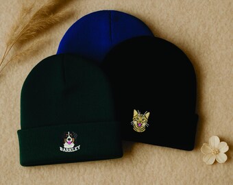 Puede incluir: Tres gorros en tonos azul, negro y verde. El gorro verde presenta un perro bordado con el nombre "BARKLEY". El gorro negro tiene una cara de gato bordada. El gorro azul es liso. Una ramita de hierba seca y una flor blanca también están en la imagen.