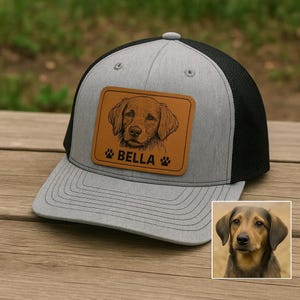 May include: A gray and black trucker hat with a brown leather patch featuring a dog portrait and the name "BELLA". The hat has a curved brim and a mesh back.