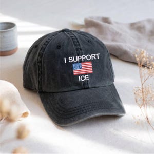 May include: A dark grey baseball cap featuring the text "I SUPPORT ICE" in white, with an embroidered American flag. The cap has a worn, distressed appearance and is made of a soft fabric.