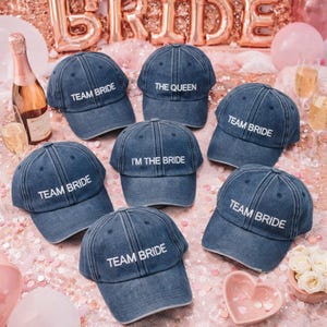 May include: A collection of denim baseball caps, perfect for a bridal party. The caps are a faded blue color and feature white embroidered text. Some caps say "TEAM BRIDE", one says "THE QUEEN", and another says "I'M THE BRIDE".