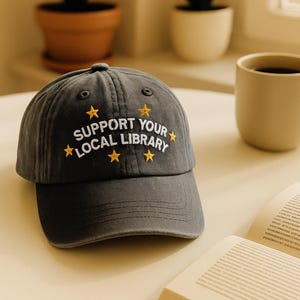 May include: A dark gray baseball cap with the embroidered text "SUPPORT YOUR LOCAL LIBRARY" in white, and yellow star accents. The cap is sitting on a light-colored surface, with a coffee mug and open book nearby.