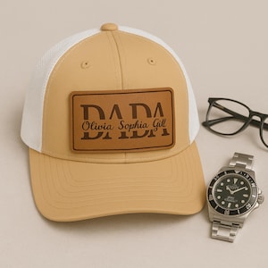 Op de afbeelding: Een bruin en witte truckerpet met een leren patch met de tekst "DADA" boven de namen "Olivia Sophia Gill". Een zilveren polshorloge en een zwarte bril staan ook op de foto.