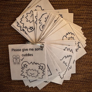 Puede incluir: Un pequeño folleto en forma de abanico con páginas blancas y un anillo de metal. Cada página tiene un dibujo lineal negro de una explosión de estrellas y otras formas. La primera página dice "Please give me some cuddles" con un dibujo de dos personas.