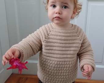 Puede incluir: Un suéter de crochet beige con cuello redondo y mangas largas. El suéter tiene un patrón de puntada texturizada y lo lleva un niño pequeño. El niño también lleva pantalones negros y sostiene una flor roja.