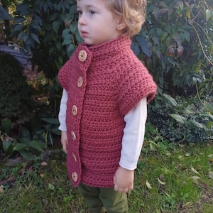 Puede incluir: Un chaleco de ganchillo burdeos con grandes botones de madera. El chaleco sin mangas tiene un cuello alto. El niño lleva una camisa blanca de manga larga, pantalones verde oliva y botas marrones. El fondo es un entorno verde y frondoso.