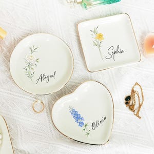 Könnte beinhalten: Drei weiße Keramik-Schmuckschalen mit goldenen Rändern. Eine runde Schale mit "Abigail", eine quadratische Schale mit "Sophia" und eine herzförmige Schale mit "Olivia". Jede Schale hat ein Blumendesign. Ein goldener Ring und eine Halskette sind ebenfalls zu sehen.