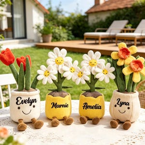 Könnte beinhalten: Vier Plüsch-Blumentöpfe mit lächelnden Gesichtern und baumelnden Beinen. Zwei Töpfe sind cremefarben mit roten Tulpen und den Namen Evelyn und Lyna. Zwei Töpfe sind gelb mit weißen Gänseblümchen und den Namen Marnie und Amelia bestickt.