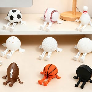 May include: A selection of sports-themed desk accessories, each designed as a ball or object with attached arms and legs. Includes a football, baseball, bowling pin, golf ball, volleyball, rugby ball, basketball, and a black disc.