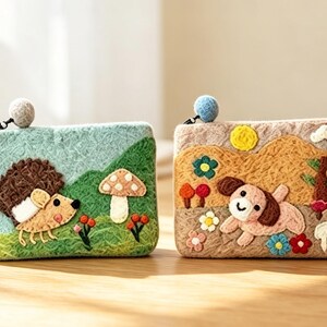Könnte beinhalten: Zwei rechteckige Filztaschen mit Reißverschluss und Bommel-Anhängern. Die linke Tasche zeigt einen Igel, grüne Hügel und einen Pilz. Die rechte Tasche zeigt einen braunen Hund, eine gelbe Sonne, Blumen und einen Baum auf beigem Grund.