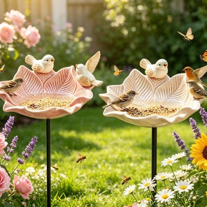 May include: Two lotus-shaped bird feeders on thin black metal stakes. One feeder is light pink, the other is off-white. Both contain birdseed and are occupied by small birds. The feeders are positioned in a garden with roses and sunflowers.