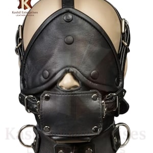 Puede incluir: Una máscara facial de cuero negro con una abertura para la nariz y una cubierta bucal rectangular. La máscara tiene correas ajustables, hebillas metálicas y un collarín con un anillo metálico. La marca "Kashif Enterprises Leather Goods" es visible.