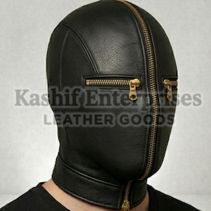 以下が含まれることがあります： 黒い革製のフード。顔全体を覆うジッパーと、目の近くにある2つの小さなジッパーが付いています。フードは頭と首を覆っています。「Kashif Enterprises LEATHER GOODS」の文字が前面に印刷されています。