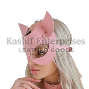 Puede incluir: Una m&aacute;scara de gato de cuero rosa con orejas puntiagudas y recortes para los ojos. La m&aacute;scara se asegura con una correa y remaches. La imagen tambi&eacute;n muestra un collar rosa a juego con un gran anillo de metal. El texto "Kashif Enterprises LEATHER GOODS" es visible.