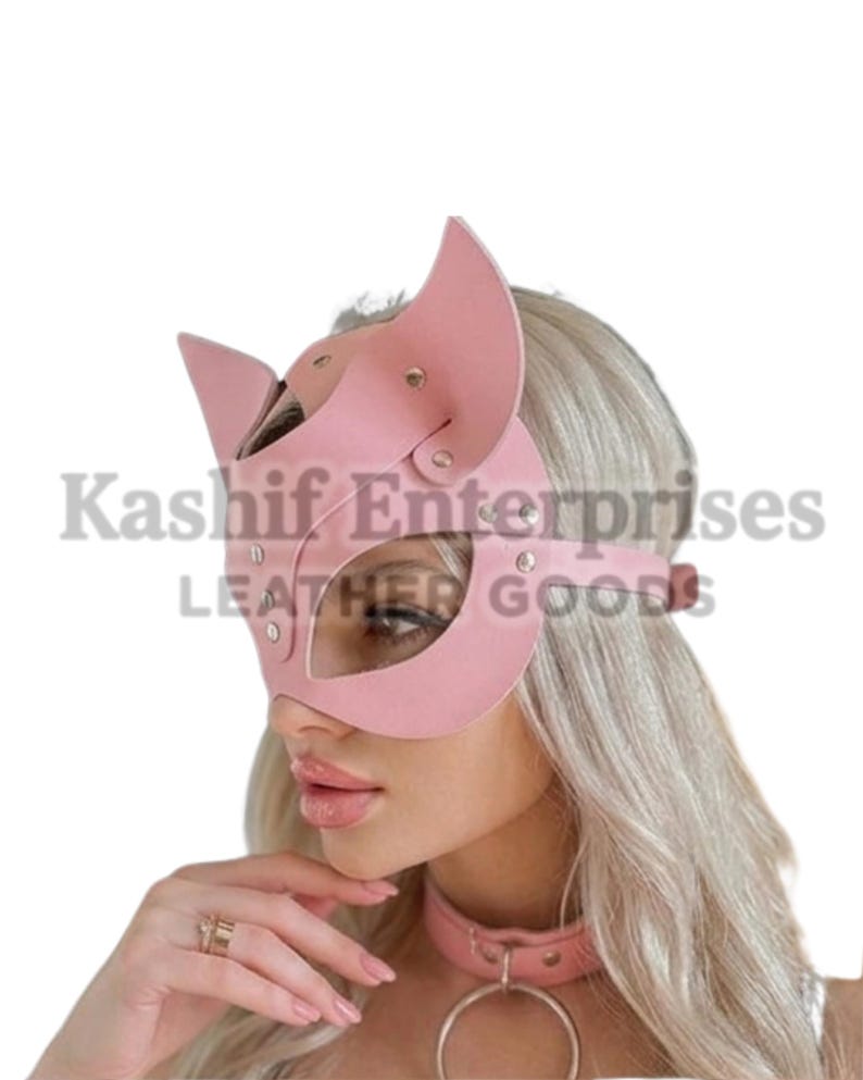 Puede incluir: Una m&aacute;scara de gato de cuero rosa con orejas puntiagudas y recortes para los ojos. La m&aacute;scara se asegura con una correa y remaches. La imagen tambi&eacute;n muestra un collar rosa a juego con un gran anillo de metal. El texto "Kashif Enterprises LEATHER GOODS" es visible.