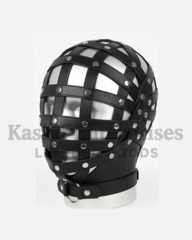 Puede incluir: M&aacute;scara de cabeza de cuero negro con un dise&ntilde;o de jaula. La m&aacute;scara presenta correas horizontales y verticales aseguradas con remaches plateados. Una correa con un anillo de metal est&aacute; alrededor del cuello. El texto "Kas" y "Goods" es visible.