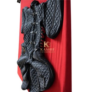 Op de afbeelding: Een zwart doorgestikt kledingstuk met een hoge kraag, meerdere verstelbare zwarte leren riemen met metalen gespen en zijpanelen geregen met rood koord. Het kledingstuk heeft lange mouwen en het gouden logo KASHIF ENTERPRISES.
