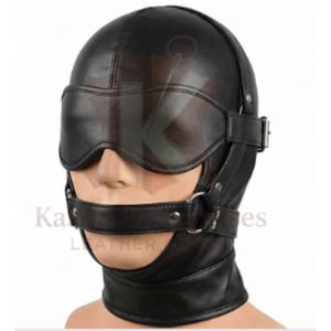Puede incluir: Capucha de cuero negro con antifaz, correa para la barbilla y pieza para el cuello. La capucha tiene un cierre de hebilla y un anillo metálico en la correa de la barbilla. El texto "Ka Leather" es parcialmente visible.