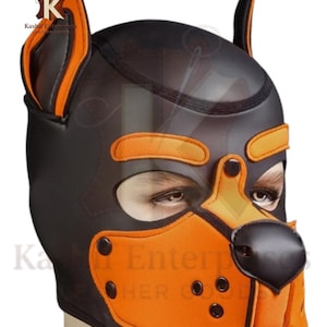 Puede incluir: Capucha de cachorro negra y naranja con diseño de perro. La capucha presenta aberturas para los ojos, una nariz y detalles de orejas. El diseño incluye detalles en naranja y remaches negros.