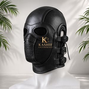 Op de afbeelding: Een zwarte lederen kap met ovale ooguitsparingen met zwart gaas, een geperforeerd neus- en mondgedeelte en drie verstelbare zijriemen met metalen gespen. De tekst KASHIF ENTERPRISES is in goud op de voorkant van het masker gedrukt.
