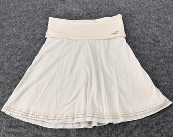 May include: A white skirt featuring a wide, fold-over waistband with a small embroidered logo on the front. The hem of the skirt is decorated with three horizontal rows of metallic silver stitching, creating a subtle, textured pattern along the bottom.