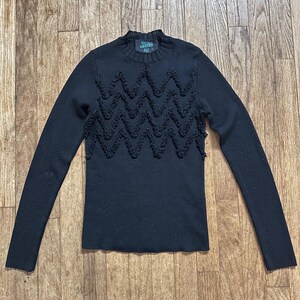 May include: Black long-sleeve sweater with a high neckline and a decorative zigzag pattern across the chest. The sweater has a ribbed texture and a small label at the neckline. The sweater is made of a soft material.