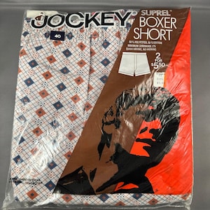 Puede incluir: Un paquete de bóxers Jockey Suprel. Los pantalones cortos tienen un fondo blanco con un patrón repetido de diamantes naranjas y azules. El embalaje incluye el logotipo de Jockey, información del producto y un gráfico de la silueta de un hombre.