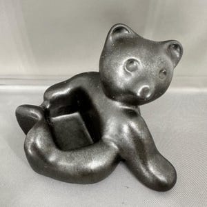 May include: A black bear figurine in a crawling pose. The bear has a smooth, glossy finish and a simple, stylized design. The figurine is made of a solid material and has a small, open area in the centre.