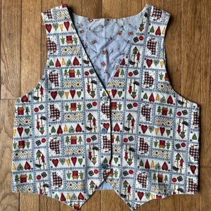 Op de afbeelding: Omkeerbaar vest met een patchworkpatroon met harten, vogelhuisjes, bomen en katten op een lichtblauwe achtergrond. Het vest heeft een V-hals en een knoopsluiting. De achterkant is een lichtblauwe stof met een klein patroon.