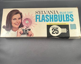 May include: Vintage Sylvania Blue Dot Flashbulbs box. The box features a woman holding a camera with a flash. The box has the words "SYLVANIA BLUE DOT FLASHBULBS" in blue and white. The box also says "PRESS 25".