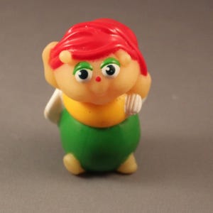 Könnte beinhalten: Vintage Kunststofffigur einer Zeichentrickfigur mit roten Haaren, grünen Augen und einem gelben Hemd. Die Figur hat einen grünen Körper und kleine weiße Flügel. Die Figur steht auf einer grauen Oberfläche.