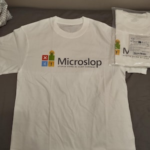 May include: White t-shirt with the text "Microslop STUPID DUMB AI SLOP COMPANY" in black and a colorful logo. The shirt is displayed on a gray surface, with a packaged shirt visible in the background.