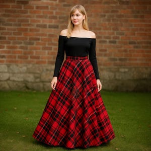 May include: A woman wearing a black off-the-shoulder long-sleeve top and a long, red and black plaid skirt. The skirt has a classic tartan pattern. The background features a brick wall and green grass.