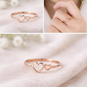 Puede incluir: Anillo de oro rosa con dos diseños en forma de corazón. El anillo se muestra sobre una tela blanca y suave, y en un dedo. El delicado diseño del anillo lo convierte en un regalo perfecto para un ser querido. Una joya elegante.