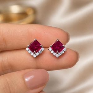 May include: A pair of stud earrings featuring a square-cut deep red gemstone at the centre, framed by a partial halo of small, clear, round-cut white stones set in a silver-toned metal base, arranged in a diamond-shaped orientation.