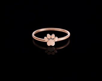 Puede incluir: Un anillo de oro rosa con un diseño de huella de pata. El anillo tiene una banda delgada y un pequeño dije detallado de huella de pata. El anillo se muestra sobre un fondo negro, resaltando su brillo metálico.