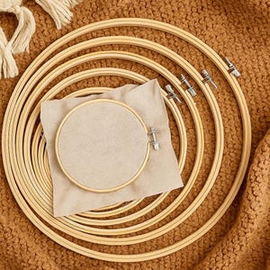 Può includere: Una collezione di telai da ricamo in legno di varie dimensioni, disposti a incastro. Un pezzo di tessuto beige è centrato, con un telaio più piccolo che lo tiene in posizione. I telai sono in legno chiaro e hanno meccanismi di serraggio in metallo.