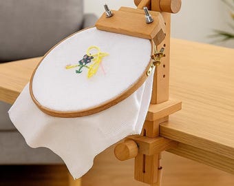 May include: A wooden embroidery hoop stand with a white fabric stretched in the hoop. The fabric has a small, colorful embroidered design. The stand is clamped to a wooden table. The stand is made of light-colored wood.