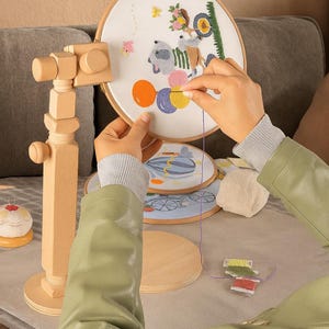 May include: An embroidery hoop with a colourful design is held in a wooden stand. The design depicts a cartoon animal on a vehicle with balloons. Other hoops and embroidery supplies are also visible.