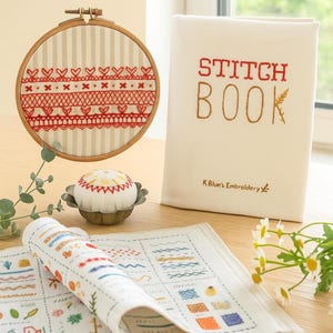Puede incluir: Un kit de bordado con un aro de madera con diseños de corazones y encajes rojos, un "Stitch Book", un alfiletero y una tela enrollada con ejemplos de puntadas. El kit está sobre una superficie de madera cerca de una ventana.