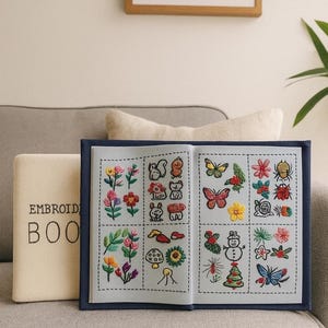 Può includere: Un libro di ricamo aperto con una copertina blu mostra colorati disegni ricamati di fiori, insetti e animali. Un libro abbinato con la scritta "EMBROIDERY BOOK" è accanto. Gli oggetti sono su un divano grigio.