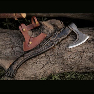 May include: A detailed, dark-toned axe with a carved handle and a silver blade, resting on a wooden surface. A brown leather sheath with brass-colored fasteners is also present. The axe features intricate dragon-like carvings.