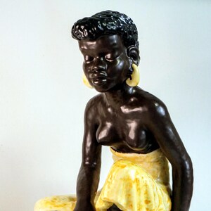 May include: A ceramic figurine of a seated woman with dark skin, wearing a yellow skirt and large yellow earrings. The figure is posed with her legs crossed and her arms resting on her legs. The figurine has a glossy finish.