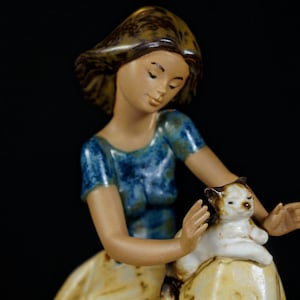 May include: A ceramic figurine of a person with brown hair wearing a blue mottled top, gently holding a small white and brown cat. The person has a serene expression, and the cat is resting on a rounded, light-colored base with brown accents.