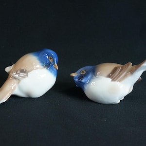 May include: Two porcelain bird ornaments. Each bird features a blue head, tan wings, and a white body. The birds are positioned facing each other, with one bird's tail extended. The ornaments are set against a black backdrop.