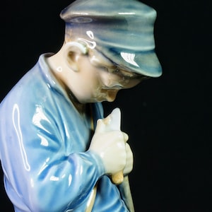 May include: A ceramic figurine of a person in a blue hat and jacket, holding a light-colored object. The figurine has a glossy finish and is set against a black background. The figure is looking down.